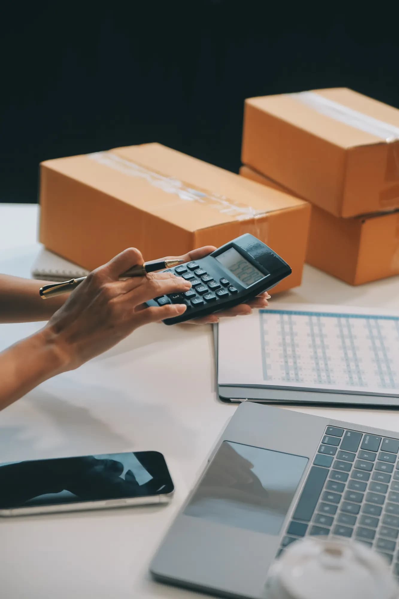 Gestora calculando preços com calculadora ao lado de caixas de mercadoria